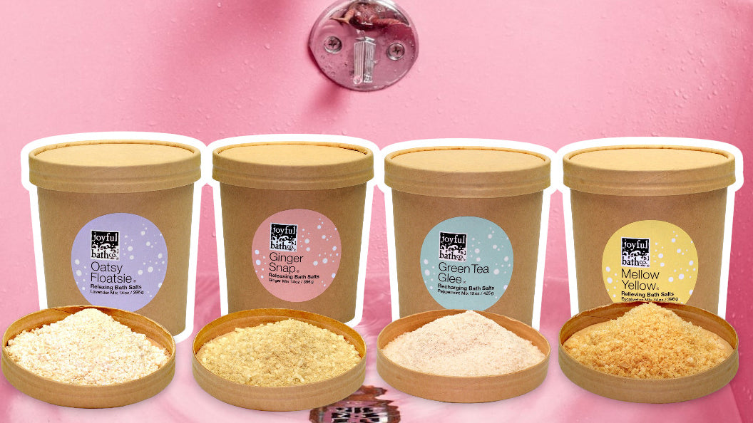 Four different bath salts in cardboard containers displayed in pink bathtub.