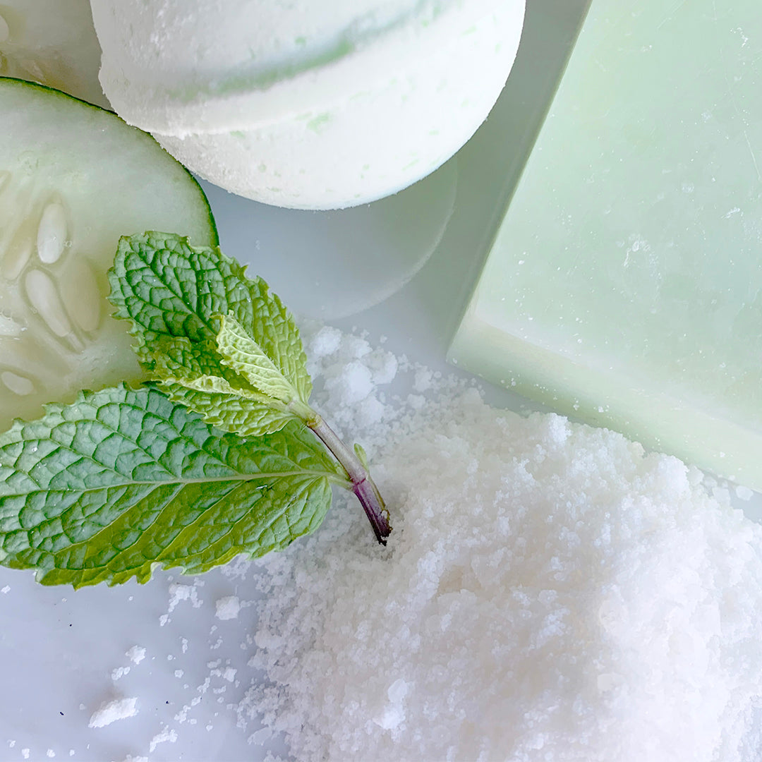 close up of veg out bath salts surrounds be other cucumber and mint plants, a light green bath bomb and soap