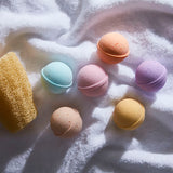 Six colorful bath bombs next to a loofah laying atop a white fluffy towel
