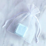 Mesh Shower Steamer Bag with drawstring pulled tight to enclose a Shower Steamer cube. Background of white marble tile.