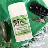 Hemp Balm on a stick laying a green towel with white tassels and next to it are a pair of sunglasses and car keys