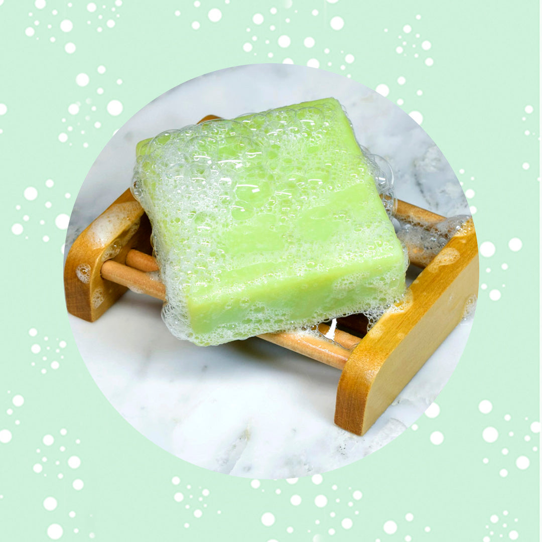 A sudsy and green cucumber mint scented  soaps sitting on a wooden soap cradle surrounded by  a green background and bubbles